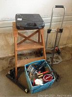 Overview of the lot showing wooden ladder, black Jobmate drill case on top, metal dolly, and blue crate with assorted yard lights and cables.