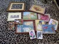 Overview of the full lot showing assortment of framed and unframed prints and paintings arranged on a patterned fabric surface. Includes a large empty frame, various framed art, and miniature paintings.