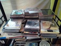 Photo showing multiple stacks of assorted CDs on a bed frame, mostly in jewel cases with visible covers of various artists and genres.
