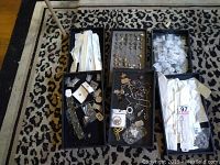 Photo showing multiple trays with jewelry pieces including rings, chains, pendants, and other items placed on a patterned rug.
