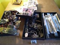 Top-down view showing black trays with mixed loose costume jewelry including necklaces and bracelets.