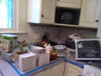 Photo showing kitchen counter with boxed Chop Magic, ceramic teapot, assorted plates and bowls, clear and amber glassware, Ronco rotisserie oven and an infrared oven appliance