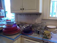 Photo showing an assortment of glassware and dishware on a counter including clear drinking glasses, red and clear glass plates, a few decorated espresso cups, and some Tupperware containers.