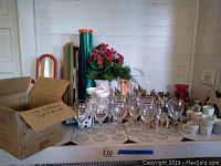 Table with 17 sets of white Crestware espresso cups and saucers, many clear stemware glasses, and a box containing more cups/saucers, along with floral arrangements in vases in the background.