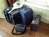 Photo showing two black hardshell suitcases, blue rolling suitcase, metal attorney's case, black Nikon camera bag, and tennis racket.