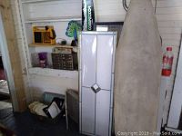 Room corner with tall white paper peacock light with diamond glass detail, yellow Beachcomber radio on shelf, fishing box/case, waterskis leaning on wall.
