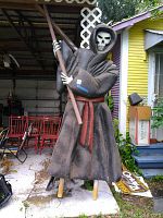 Full view of large grim reaper figured molded in polystyrene, standing on wooden legs, with black cloak, skeletal face, and wooden scythe staff.