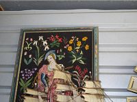 Framed tapestry showing a woman in pink and blue with flowers on a dark background, partially obscured by a model ship.