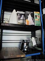 Overview of the lot on black shelving, showing boxes of Rowenta iron, Prime Time coffee carafe, Presto Salad Shooter, white lidded pan, wood cutting board and Osterizer blender base with glass jar.