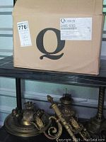 Photo of a cardboard box labeled Quorum International containing a charcoal colored chandelier. Below the shelf are brass colored light fixtures and parts including what appears to be the base of a hurricane oil lamp without the glass shade.
