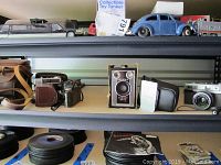 Shelf with multiple vintage and digital cameras including Brownie, Minolta, Olympus SZ11, and others.