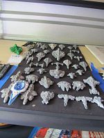 Top-down view showing a large assortment of miniature metal sci-fi spaceships laid out on a blue surface, mostly unpainted metal, varying shapes and sizes with detailed designs. One spaceship painted blue and white.