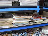 Stack of comic books on a shelf, some in plastic sleeves, showing a variety of titles and sizes.