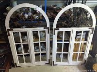 Two white wooden wall hanging window frames with metal decorative arched tops, placed on the floor leaning against blue metal shelving with dishes in the background.