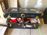 Black metal foot locker trunk open with holiday decor inside, showing wear and brass corner protectors