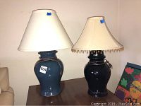 Two ceramic base lamps against a plain wall; the taller blue lamp has a plain white shade, the shorter black lamp has a beaded fringe shade.