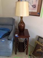 Full view of wood table with magazine rack and brown ceramic lamp with shade on top, next to blue armchair and framed print on wall.