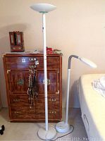Two white floor lamps next to wooden cabinet and bed, showing their shape and size