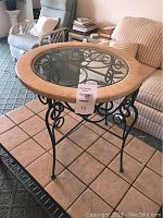 Front view of a round end table with a glass top and green iron base showing decorative scrollwork and some scratches on the frame.