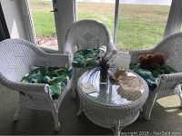 Set of three white wicker chairs with tropical green leaf cushions and a round white wicker coffee table with glass top in a sunroom.