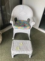 Full view of white wicker rocking chair with multi-colored cushion and matching wicker ottoman placed on green carpet.