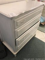 View of white chest of drawers with open lower drawer missing a handle.