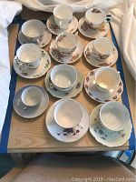 Full view of the 11 tea cups and 11 saucers arranged on a table with various floral designs in bone china, showing gold detailing.