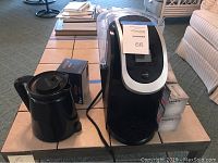 Keurig 2.0 coffee maker and black coffee carafe on table with boxes of filters and coffee pods.