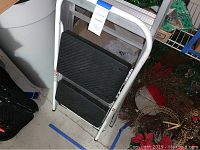 Photo showing white metal two-step stool with black tread steps leaning against wall and nearby items