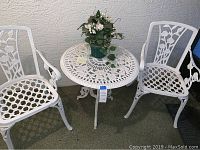 White cast iron round bistro table with floral openwork pattern and two matching chairs with floral backrests and lattice seats, all painted white, set against a textured wall.