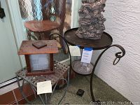 Photo showing the 25-inch round metal plant stand with metal scrolls and the dark cherub statue on top, plus the 9.5-inch square metal plant stand with wooden birdhouse on it.
