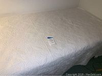 Twin mattress covered in white quilted bedspread with visible tag on top, showing item number.