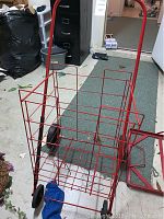 Photo showing red metal wire shopping caddy on wheels with handle in a room with carpet and tile floor.