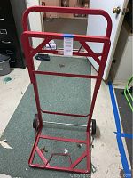 Photo showing red metal recycle bin cart frame with tubular handle and two wheels on a carpeted floor, designed to hold two recycle bins.