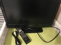 Front view of Isymphony LC19IH56 19-inch LCD TV with remote control, power cable, and AV composite cable on a green surface.