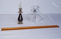 Two vintage perfume bottles and wooden ruler for scale, including dark glass bottle with brass base and teardrop stopper, and clear crystal olec cassini bottle