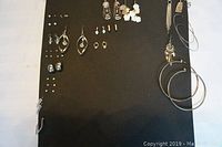 Front view of black display board showing multiple pairs of assorted costume jewelry earrings pinned on it, including silver hoops, faux pearl studs, and dangling earrings with floral designs.