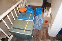 Photo showing two sleeping bags in blue storage bags, two coolers (one large green Koolatron electric cooler and one blue cooler), an orange camping lantern, and a denim or grey camping blanket. Several campfire roasting sticks are also visible in box.