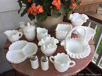 Photo of 14 pieces of white hobnail milk glass items including various bowls, baskets, and shakers arranged on a wooden table with a background flower pot.