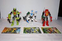 Image shows three Lego Hero Factory figures Breez 2.0 (green), Stormer 3.0 (white), Furno 3.0 (red) standing in a row with their original instruction booklets laid out in front of them on a flat surface.