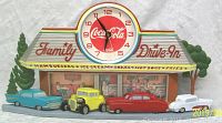 Front view showing full clock with Coca-Cola logo on clock face, colorful diner roof, four vintage cars, and background diner scene along with trees.