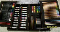 Image showing open wooden art supply case displaying colored pencils, markers, and acrylic paint tubes in compartments.