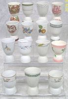 Front view of all 15 large size vintage egg cups displayed on a clear stepped stand showing various painted and embossed designs