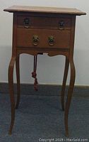 Front view of antique 2 drawer sewing table showing curved legs and metal ring pulls on drawers with burgundy tassel hanging below.
