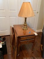 Wood end table with two drawers, brass handles, and curved legs with brass lamp on top. Two wooden folding trays are leaning against the table.
