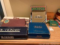 Photo of multiple boxed board and card games including Clue, Pictionary, Jackpot, Scruples and Trivial Pursuit arranged on a table against wall.