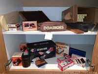 Shelf with boxed playing cards, dealer shoe, dominoes in brown case, poker chips, racks, dice cups, cribbage boards.