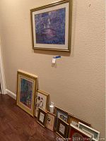 Seven framed prints arranged leaning against a wall, largest is abstract figure bridge scene in gold frame, other smaller prints with botanical and house motifs.