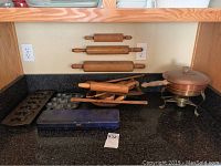 Image showing four wooden rolling pins mounted horizontally on wall, metal baking molds on counter, blue knife storage case, and copper chafing pot on brass stand.