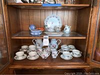 Display of Bavaria tea cups, saucers, cream and sugar, platter, and Wedgwood trinket holder and vase on wooden shelves.